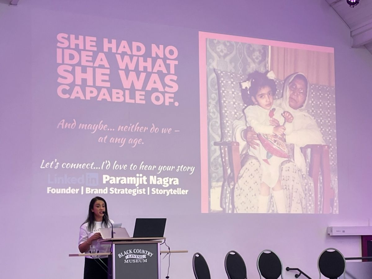 An image of Paramjit Nagra, at the Black Country Living Museum's stage, standing behind a podium, behind her is a projected presentation with a childhood photo of her with her on the right half of the screen. Her LinkedIn details, job title located on the bottom left corner. It also includes text in the top right corner saying "She had no idea what she was capable of. And maybe... neither do we - at any age"